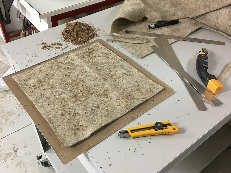 Zuschnitt eines quadratischen Naturstofftextils. Darauf gestreut sind kleine Schnipsel eines grasänlichen Gewächses.